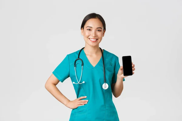 Covid-19, trabajadores de la salud y concepto de medicina en línea. Profesional médico asiático mujer, enfermera en matorrales mostrando la pantalla del teléfono inteligente y sonriendo, mostrando aplicación cita en Internet, fondo blanco — Foto de Stock