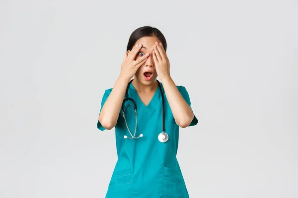 Covid-19, pracownicy służby zdrowia i zapobieganie koncepcji wirusa. Przerażona i zszokowana azjatycka pielęgniarka, lekarz w fartuchu świadkiem czegoś żenującego lub przerażającego, zakryć oczy, ale zerknąć przez palce — Zdjęcie stockowe