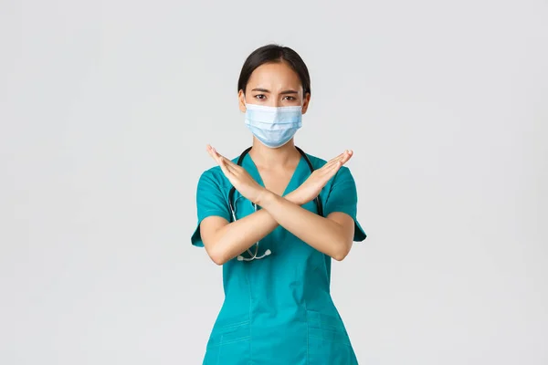 Covid-19, coronavirus ziekte, gezondheidswerkers concept. Ernstig ontevreden, fronsende aziatische arts in scrubs tonen kruis gebaar in afkeuring, iemand stoppen, verbieden actie, witte achtergrond — Stockfoto