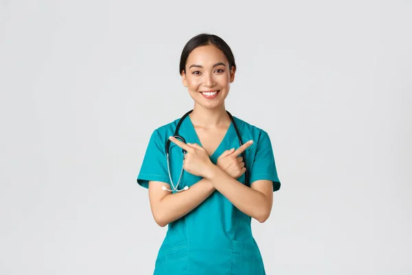 Covid-19, healthcare workers, pandemic concept. Smiling beautiful asian nurse, intern or doctor in scrubs pointing sideways and looking at camera, showing variants, two choices, white background — Stock Photo, Image