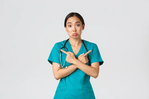 Covid-19, healthcare workers, pandemic concept. Indecisive and clueless asian female nurse, intern dont know what choose, pointing fingers sideways and shrugging unaware, white background