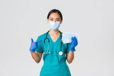 Covid-19, koronavirüs hastalığı, sağlık çalışanları konsepti. Gülümseyen Asyalı doktor, doktor veya hemşire kauçuk eldivenli ve önlüklü hasta güvenliği için tıbbi maskeleri işaret ediyor.