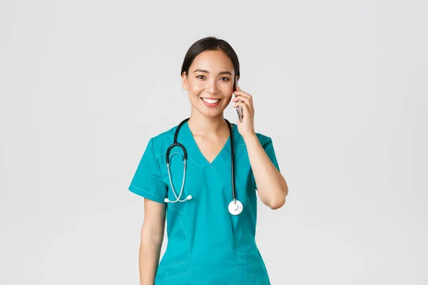 Covid-19, zdravotničtí pracovníci a prevence viru. Usmívající se šťastný asijské žena lékař, mladý internista v drhnutí mluví na telefonu, při pohledu na fotoaparátu nadějné, bílé pozadí — Stock fotografie