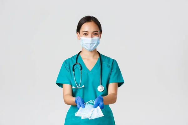 Covid-19, choroba koronawirusowa, koncepcja pracowników służby zdrowia. Przyjazny uśmiech azjatycki lekarz, kobieta lekarz w fartuchu i gumowe rękawiczki przekazujące maski medyczne pacjentom, białe tło — Zdjęcie stockowe
