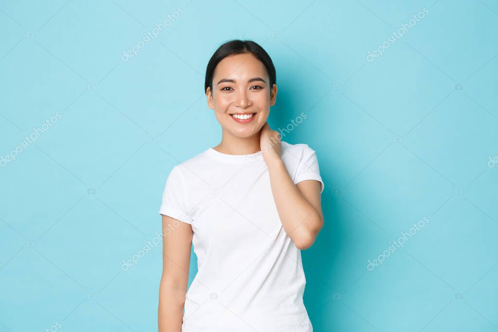 Portrait of beautiful modest smiling asian girl looking happy at camera, grinning and touching neck shy, blushing over compliment, standing satisfied over light-blue background