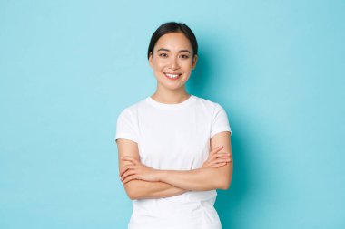 Kendine güvenen Asyalı kızın portresi. Memnun, kolları çapraz, kendine güvenen duruş, neşeli ve kararlı görünen bir kız öğrenci. Mavi arka plan, günlük kıyafetler, yaşam tarzı konsepti.