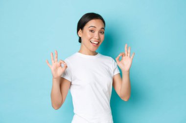 Asyalı gülümseyen kız her şeyi kontrol altında tutuyor. Kadınlar her türlü iyi davranışı sergilemeyi teşvik eder. Onaylama, kalite garantisi, tatmin olmuş mavi arka plan.