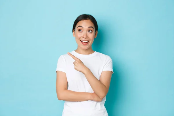 Estilo de vida, belleza y concepto de compras. Emocionado y divertido hermosa chica asiática en blanco camiseta apuntando esquina superior izquierda y sonriendo emocionado con publicidad impresionante promo, fondo azul —  Fotos de Stock