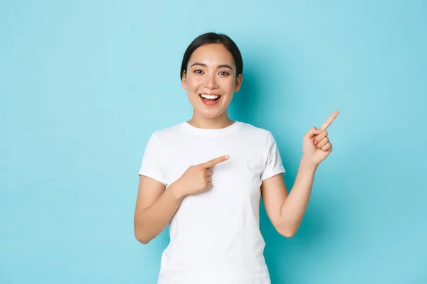 Прекрасна щаслива азійська дівчина в білій футболці допомагає з вибором, показує шлях або демонструє прапор. Корейська жінка з ентузіазмом вказує на верхній правий кут над синім фоном. — стокове фото