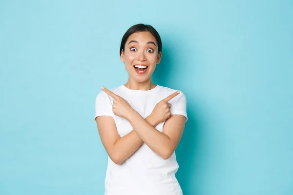 Sorprendido chica asiática feliz y sorprendida haciendo anuncio, mujer señalando los dedos hacia los lados y espacios vacíos para su bandera, mirando asombrado y alegre, mostrando el camino, fondo azul —  Fotos de Stock