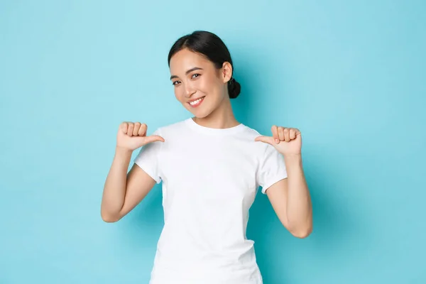 Fiduciosa donna asiatica sorridente in t-shirt bianca che si indica con espressione orgogliosa e assertiva, esibizionista, promuove le proprie capacità, cerca lavoro, in piedi sfondo blu — Foto Stock