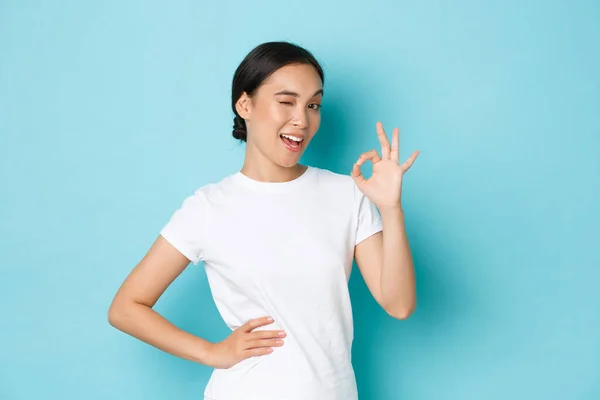 Retrato de atrevida e confiante muito asiático menina 20s em branco t-shirt, garantir tudo sob controle, mostrando ok gesto, aprovar ou como produto, piscando incentivando, fundo azul — Fotografia de Stock