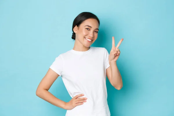 Lindo sonriente asiático hembra en blanco camiseta, mostrando paz kawaii signo y buscando adorable, coqueteando, permaneciendo positivo o optimista, de pie azul fondo feliz actitud —  Fotos de Stock