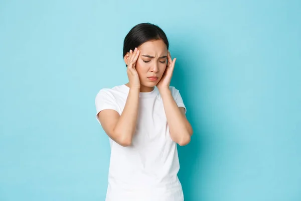 Vista lateral de molestado chica asiática cansada haciendo muecas, tocando la cabeza y quejándose de dolor de cabeza, tener dolor en la cabeza, migraña de sentirse mareado, de pie fondo azul enfermo — Foto de Stock