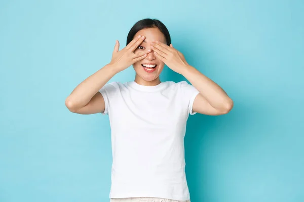 Retrato de alegre hermosa chica asiática mirando a través de los dedos mientras se cierran los ojos con las manos, sonriendo optimista, curioso lo que sucede alrededor, de pie fondo azul — Foto de Stock