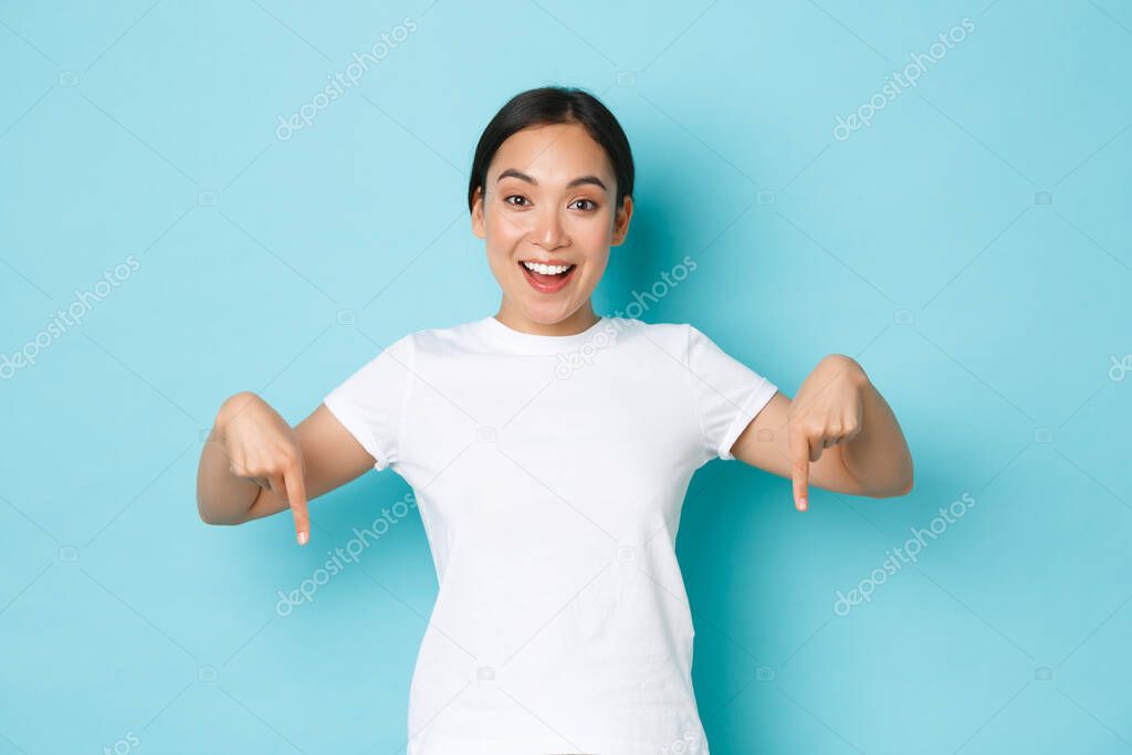 Cheerful young asian lady in white t-shirt pointing fingers down and smiling excited, looking upbeat while demontrating banner, offer special discount promo, standing blue background
