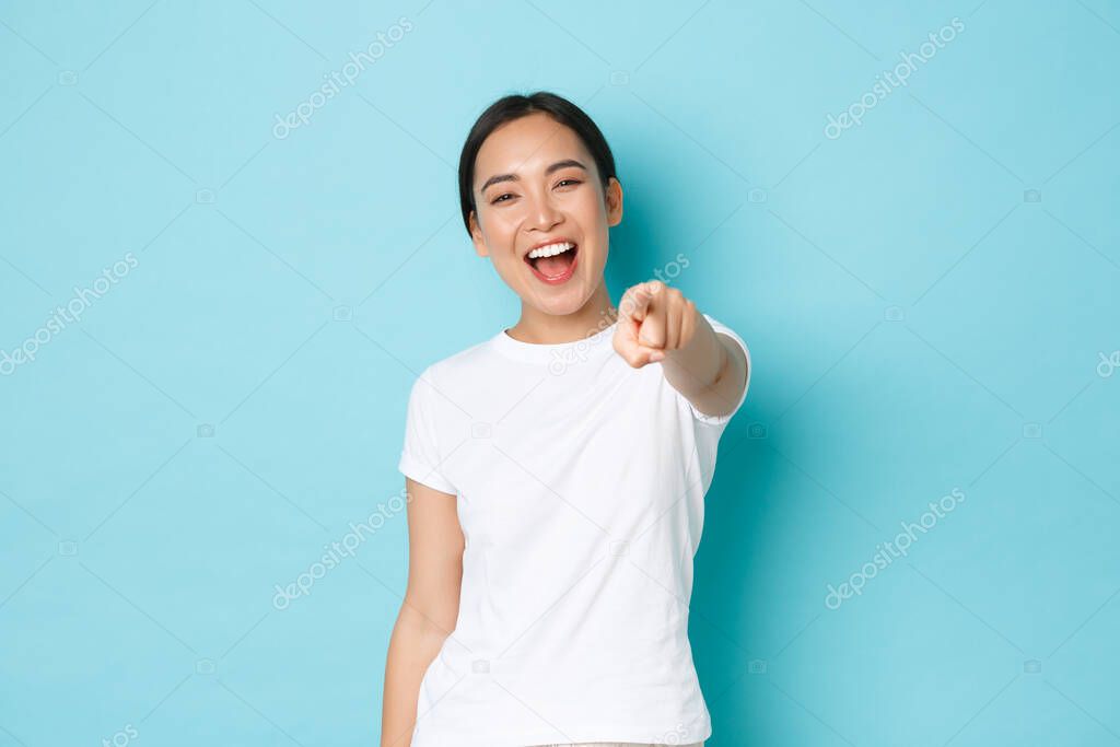 Carefree upbeat pretty asian girl in white t-shirt having fun, laughing and pointing finger as if mocking person, having fun of hilarious friend, standing happy over blue background