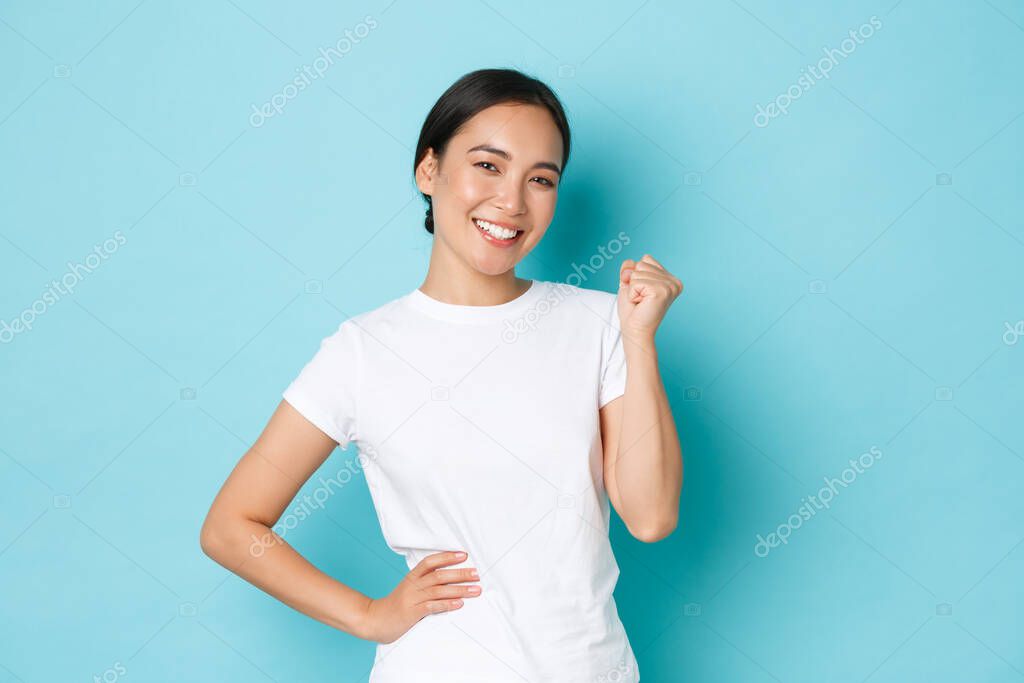 Successful winning asian girl in white t-shirt rejoicing over small victory, clench fist satisfied, triumphing, smiling pleased and sassy, become champion, standing blue background