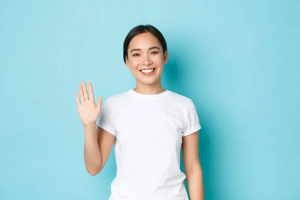 Retrato de amigable chica asiática alegre en camiseta blanca diciendo hola, saludando a la gente, sonriendo y saludando la mano en un gesto de alta, de pie fondo azul optimista, bienvenida a alguien —  Fotos de Stock