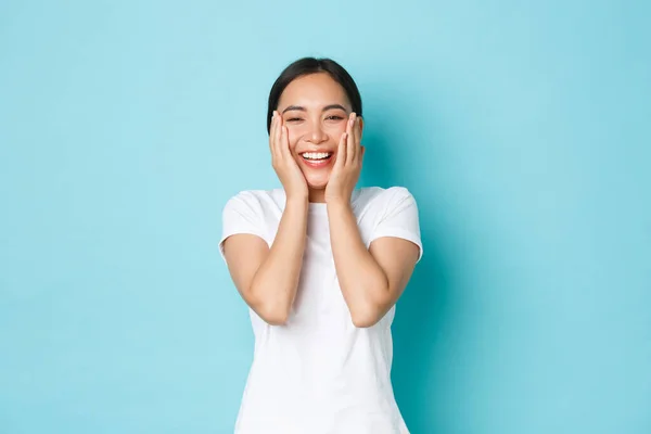 Skincare, keindahan dan konsep gaya hidup. Gadis asia yang ceria tersenyum bersukacita, terlihat bahagia, menyentuh kulit yang bersih dan bersukacita, menyingkirkan jerawat, latar belakang biru berdiri — Stok Foto