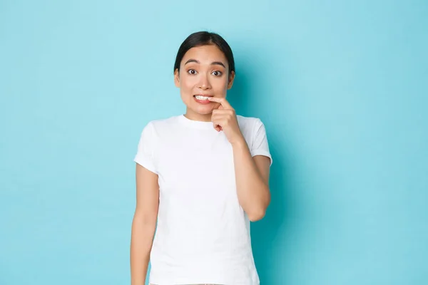 Besluiteloos schattig en dom aziatisch meisje bijten vinger en kijken aarzelend, te bang om riskante vraag te stellen, staan bedachtzaam en twijfelachtig over blauwe achtergrond in casual wit t-shirt — Stockfoto