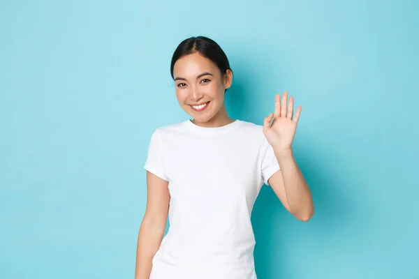 Amable linda y tímida chica asiática en blanco casual camiseta diciendo hola, sonriendo tonta y saludando la mano en gesto de hola, de pie optimista sobre fondo azul, persona de saludo —  Fotos de Stock