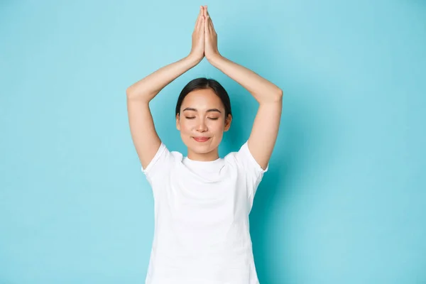 Porträtt av glad och fridfull, avslappnad leende asiatisk flicka som tar hand om sin mentala hälsa, utöva yoga eller meditation, blunda och höja händerna ovanför huvudet i asana pose, blå bakgrund — Stockfoto