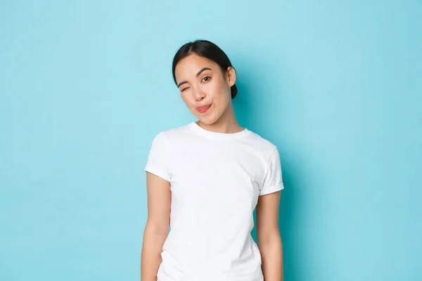 흰색 캐주얼 티셔츠를 입은 쾌활하고 매력적 인 동양 소녀의 모습, 혀를 보여 주고 윙크하는 코 쿼 티쉬, 파란 배경 위에 서서 카메라를 바라보는 대담 한 모습 — 스톡 사진