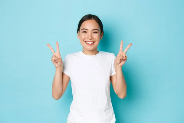 Güzellik, moda ve yaşam tarzı konsepti. Beyaz tişörtlü güzel Asyalı kızın portresi mavi arka planda duruyor, gülümsüyor ve Kawai barış jestini gösteriyor, pozitiflik ve neşe yolluyor. — Stok fotoğraf