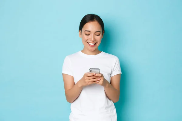 Livsstil, teknik och e-handel koncept. Porträtt av söt asiatisk flicka i vit casual t-shirt ler mot mobiltelefon skärmen. Koreanska kvinna med hjälp av smartphone ansökan över blå bakgrund — Stockfoto