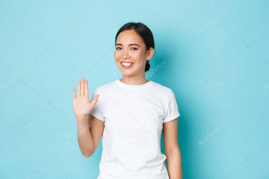 Smiling friendly-looking asian girl in white t-shirt waving hand in hello gesture, saying hi, greeting person and standing blue background happy, glad to meet someone