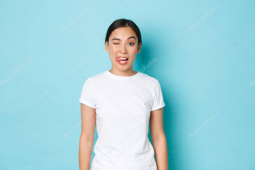 Cheeky asian girl in white t-shirt sending positive vibes, enjoying summer, showing tongue, winking carefree, standing blue background upbeat. Korean female student express joy and positive attitude
