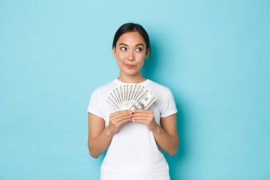 Alışveriş, para ve finans konsepti. Düşünceli ve yaratıcı güzel Asyalı kız parayı nereye yatıracağını düşünüyor, parayı tutuyor, sağ üst köşeye bakıyor, seçim yapıyor, ne alacağına karar veriyor.
