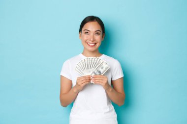 Alışveriş, para ve finans konsepti. Hayalperest gülümseyen güzel Asyalı kız parasıyla ne alacağını düşünüyor, memnun görünüyor, siparişi için ödeme hazırlıyor, açık mavi arka planda duruyor, heyecanlı.