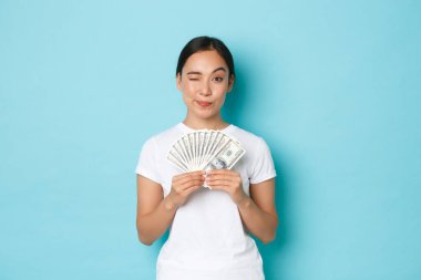 Alışveriş, para ve finans konsepti. Beyaz tişörtlü başarılı bir Asyalı kız, elinde nakit para, sana gülümseyen ve göz kırpan, yurt dışında çalışma programları için şirket öneriyor.