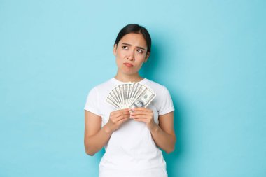 Alışveriş, para ve finans konsepti. Düşünceli ve yaratıcı güzel Asyalı kız parayı nereye yatıracağını düşünüyor, parayı tutuyor, sağ üst köşeye bakıyor, seçim yapıyor, ne alacağına karar veriyor.
