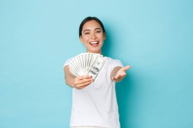 Alışveriş, para ve finans konsepti. Samimi ve nazik güzel Asyalı kız sana para veriyor. Dolarlarla dolu bir hediye, kameraya bakıyor ve gülümsüyor. Açık mavi arka plan.