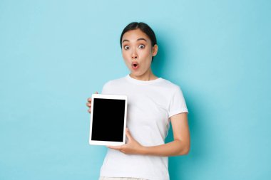 Beyaz tişörtlü, heyecanlı ve şaşkın Asyalı kızın portresi, dijital tablet ekranda harika bir şey gösteriyor, proje veya alışveriş uygulaması gösteriyor, açık mavi arka plan