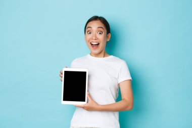 Beyaz tişörtlü, dijital tablet ekranı gösteren ve ağzı açık olan Asyalı kız hayran kaldı. Uygulama ilanı veya alışveriş sitesi, mavi arka plan.