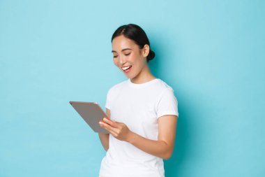 Gülümseyen güzel Asyalı kız, üç boyutlu kadın sanatçı veya serbest çalışan dijital tablet kullanan ve cihaz ekranına, online alışverişe ya da açık mavi arka planda gezinmeye bakan kişinin portresi