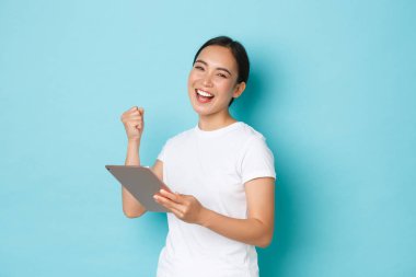 Başarılı bir şekilde gülümseyen Asyalı serbest çalışan, Dijital tablet ve yumruk pompası kullanan kız sevinç, zafer, iyi anlaşma veya proje, kutlama, açık mavi arka plan