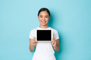 Gülümseyen güzel Asyalı bir kadının portresi dijital tablet ekranını memnun ifadesiyle gösteriyor, alışveriş sitesini gösteriyor, sanatçı için başvuru yapıyor, açık mavi arka planda duruyor.