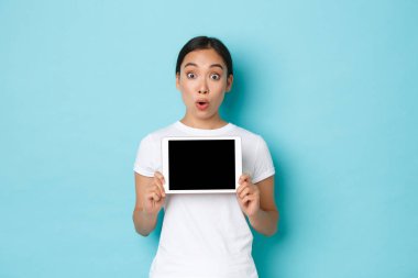 Heyecanlı ve şaşırmış Asyalı kızın portresi şaşırmış ve merak etmiş, dijital tablet ekranı gösteren ve kameraya bakan, açık mavi arka plan