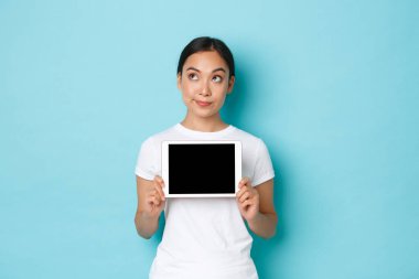 Düşünceli, düşünceli, güzel bir Asyalı kadının portresi sol üst köşeye bakarken dijital tablet ekranda belirsiz bir şey gösteriyor açık mavi arka planda.