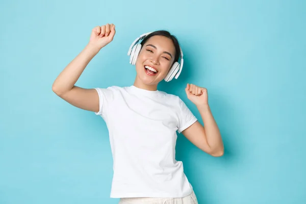 Shopping, money and finance concept. Upbeat asian girl in white casual t-shirt, singing karaoke, playing music app, listening music in wireless earphones and enjoying perfect sound