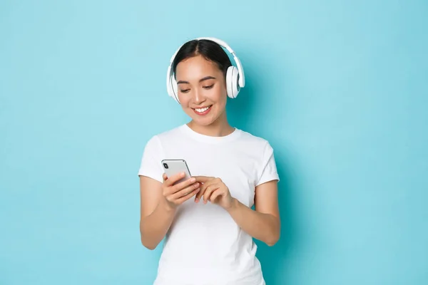 Estilo de vida, personas y concepto de ocio. Sonriendo atractiva hembra asiática usando auriculares inalámbricos y teléfono móvil, viendo video en un lugar lleno de gente, escuchando música, fondo azul claro — Foto de Stock