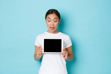 Heyecanlı ve şaşırmış beyaz tişörtlü güzel Asyalı kadın dijital tablet ekrana bakıyor, şaşırmış, heyecanlı bir şekilde bakıyor, grafiği gösteriyor ya da online alışveriş sitesini gösteriyor, açık mavi arka plan