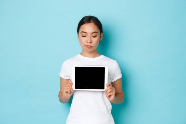 Şüpheci ve kasvetli Asyalı kız tedirgin ya da şüpheli görünüyor, dijital tablet ekranı gösteriyor, kafası karışmış bir ifadeyle, kötü ya da karmaşık bir şey gösteriyor, açık mavi arka plan