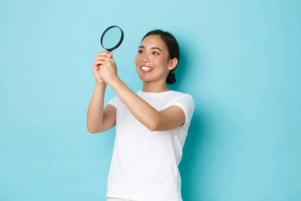 Leende entusiastisk ganska asiatisk flicka forska, utforska omgivningen, tittar genom förstoringsglas som om studera eller söka något, stående optimistisk över ljusblå bakgrund — Stockfoto