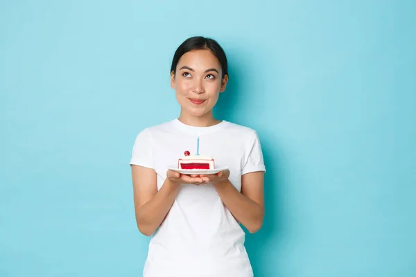 Vacaciones, estilo de vida y concepto de celebración. Retrato de niña asiática sonriente de ensueño en camiseta blanca mirando hacia arriba reflexivo mientras sostiene pedazo de pastel de cumpleaños, pidiendo deseo en vela encendida — Foto de Stock